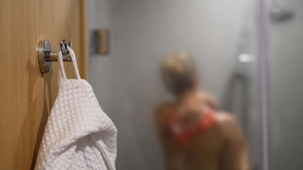 White bathrobe on hook and woman in shower in background. Morning and evening shower and body hygiene concept