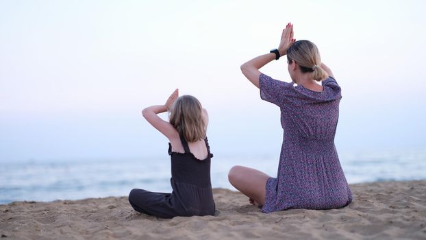 Mother and daughter do yoga meditate in lotus position on beach. Family yoga class by sea concept