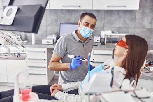 Professional male doctor uses ultraviolet lamp after making a filling on a tooth for joyful happy caucasian woman in modern dental cabinet