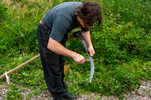 A man mows the grass with a hand scythe. Mow the grass with an old scythe. The farmer traditionally mows the lawn. The mower mows the green grass with a manual metal scythe.