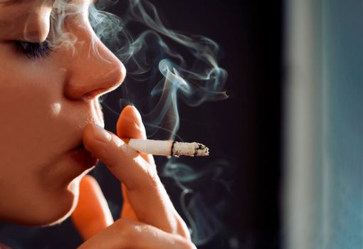 A young beauty girl is smoking, a woman is stressed and holds a cigarette in her hands, staying near the window of her apartment, close-up view.