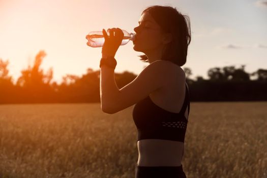 A young girl takes a break during an intense workout and jogging and drinks refreshing cool water from a bottle, a woman trains outdoor in the evening at sunset.