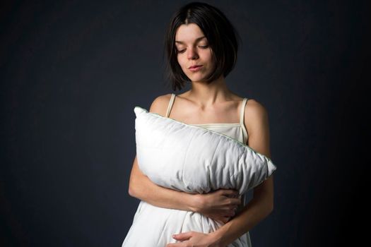 A young girl stands and hugs a white pillow on a gray background, a woman wants to sleep, take care of her health, the quality of sleep.