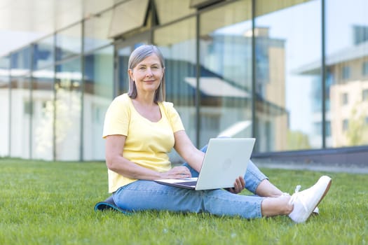 Senior happy gray-haired woman sitting on the grass and looking at the camera, working on a laptop outside the office