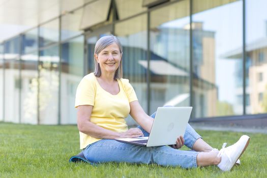 Senior happy gray-haired woman sitting on the grass and looking at the camera, working on a laptop outside the office