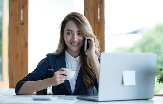 Happy young Asian business woman sitting smiling and talking on mobile phone in office..