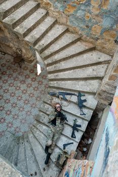 Caucasian woman in military uniform lies on the stairs of an abandoned building and holds a machine gun. View from above