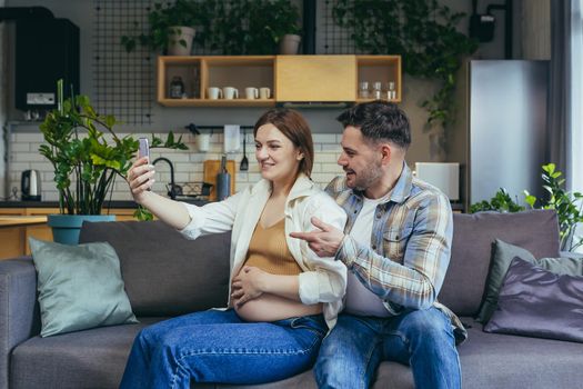 Happy young married couple man and pregnant woman using phone for video call, hugging and sitting on sofa smiling and looking at smartphone screen
