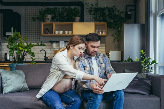 Online shopping. Young family, pregnant woman and man make an online purchase order from a laptop, holding a credit card. Sitting at home on the couch. Very happy, satisfied.