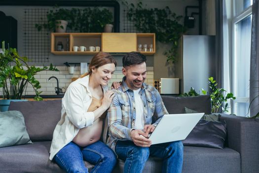 Young married couple man and pregnant woman at home on sofa using laptop for online shopping and video communication