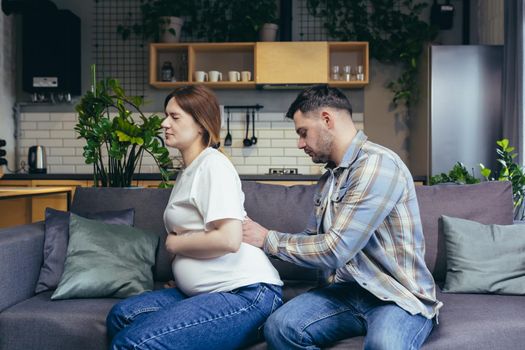 Young family. Husband massages the back of his pregnant wife. Sitting on the couch. The woman is in pain. In anticipation of childbirth