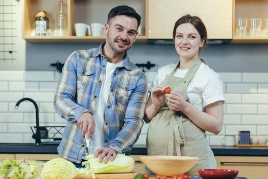 Portrait of young happy family man and pregnant woman together in the kitchen preparing healthy food, rejoicing and smiling looking at camera