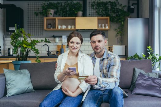 Young married couple husband and pregnant woman sitting together on the couch and hugging using the phone for online shopping in the online store, holding a bank credit card