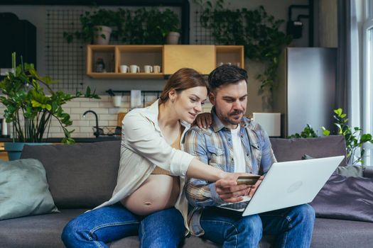 Young married couple man and pregnant woman at home on sofa using laptop for online shopping and video communication