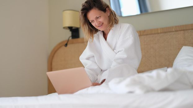 Woman sits at home on bed in white bathrobe and works on laptop. Work from home concept of quarantine and remote work freelancing