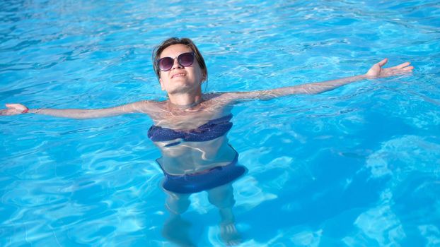 Charming woman in sunglasses enjoys swimming in turquoise water of pool. Relax in luxury hotel pool and wellness concept