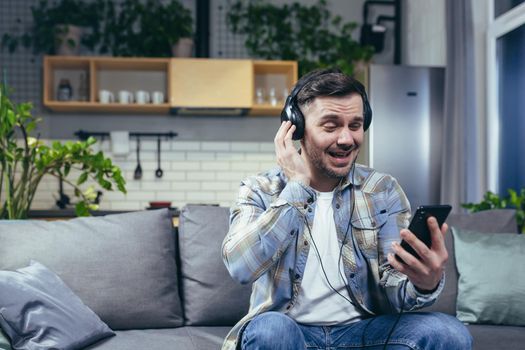 Happy man at home smiling and listening to music from phone uses music app on smartphone online, with big headphones