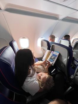 Mom with a sleeping daughter in her arms sits in an airplane seat and reads a magazine. High quality photo