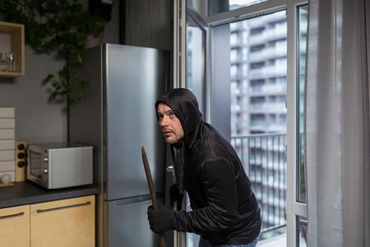 Young male robber in dark clothes carefully and quietly enters through the balcony window into the apartment, house. Holds scrap metal, fittings.
