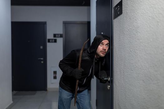 A young male robber in dark clothes stands carefully and quietly at the door of the apartment in the entrance. He listens to see if anyone is at home, checks the door handle. Holds scrap, fittings. He breaks down the door