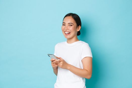 Lifestyle, technology and e-commerce concept. Side view of attractive asian girl using mobile phone, texting, messaging or chatting with friends online, starting to laugh and look at camera.