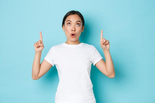 Surprised and impressed asian girl in white t-shirt looking and pointing fingers up at something interesting and amazing. Beautiful korean girl 20s found awesome promo offer, over blue background.