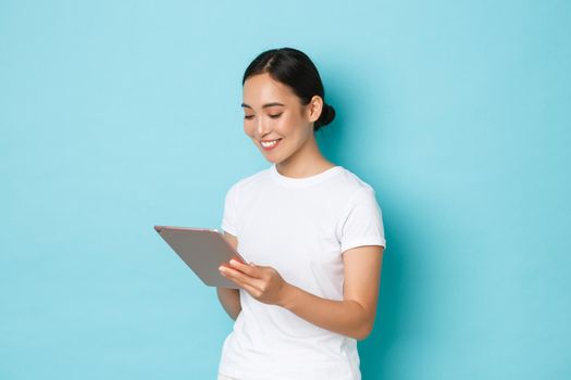 Portrait of smiling beautiful asian girl, female 3d artist or freelancer using digital tablet and looking pleased at device screen, shopping online or browsing over light blue background.