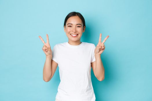 Beauty, fashion and lifestyle concept. Portrait of beautiful asian girl in white t-shirt standing over blue background, smiling and showing kawaii peace gesture, sending positivity and joy.