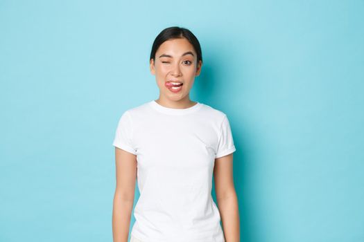 Cheeky asian girl in white t-shirt sending positive vibes, enjoying summer, showing tongue, winking carefree, standing blue background upbeat. Korean female student express joy and positive attitude.