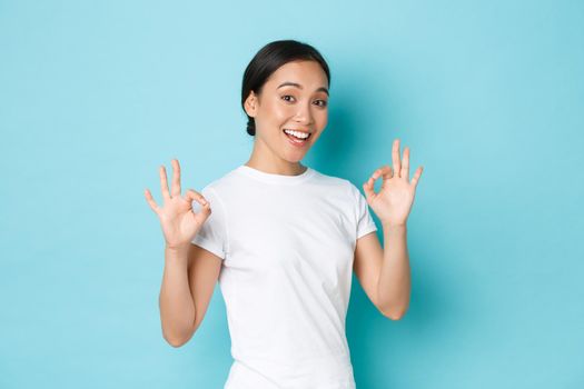 Pretty confident asian smiling girl ensure everything OK, under control. Woman encourage all good, showing okay gesture and nod in approval, guarantee quality, standing satisfied blue background.
