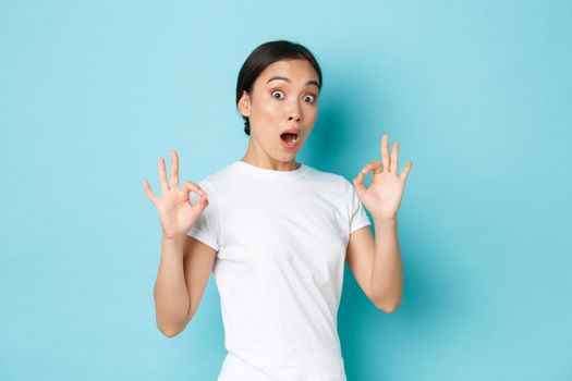 Excited and impressed asian girl in white t-shirt look surprised and astonished with amazing, perfect service, showing okay gesture and looked astounded over blue background.
