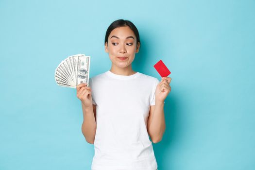 Shopping, money and finance concept. Dreamy beautiful asian girl looking happy and thoughtful upper left corner, smiling while choosing what buy on cash and with credit card.