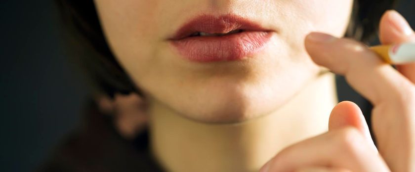 A young beauty girl is smoking, a woman is stressed and holds a cigarette in her hands on the dark background, close-up view of her face, lips.