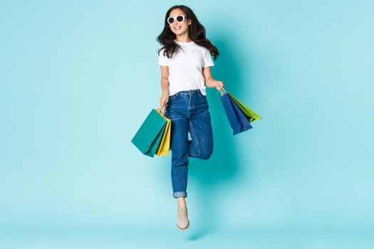 Fashion, beauty and lifestyle concept. Attractive carefree asian woman in sunglasses, holding shopping bags and smiling delighted, enjoying walking in mall, jumping upbeat over light blue background.