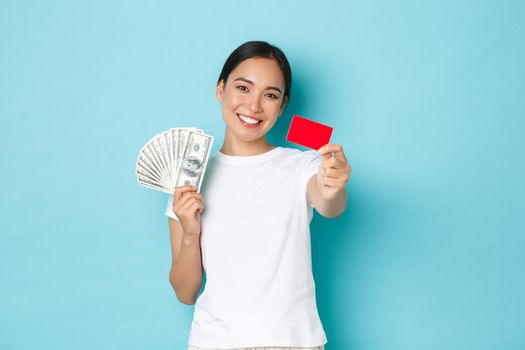 Shopping, money and finance concept. Happy carefree asian girl in white t-shirt holding cash but choosing credit card, like contactless payment, smiling upbeat, blue background.