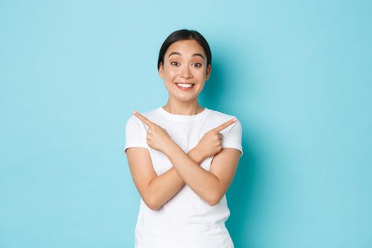 Cheerful, happy and excited asian girl in white t-shirt, pointing fingers sideways, showing left and right variants, demonstrate choices for you, smiling upbeat over blue background.