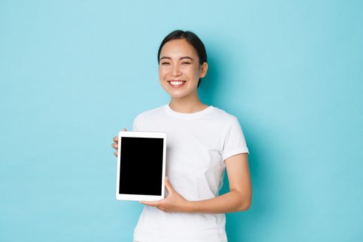 Portrait of proud and happy smiling asian girl, grinning joyfully, looking pleased while showing digital tablet screen, demonstrate her work project or advertisement, light blue background.