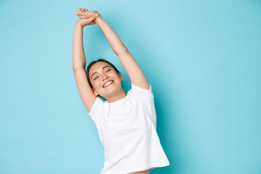 Portrait of carefree and relaxed, beautiful asian girl close eyes and stretching hands upwards with happy smile, feeling fresh and cheerful after taking nap, standing blue background.