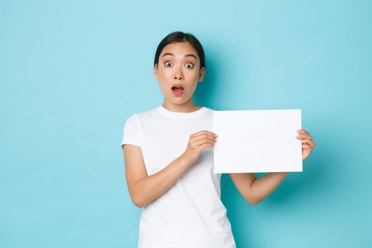 Amazed and excited asian girl in white t-shirt, making announcement, gasping surprised while showing sign, piece of blank paper with your advertisement, standing light blue background.