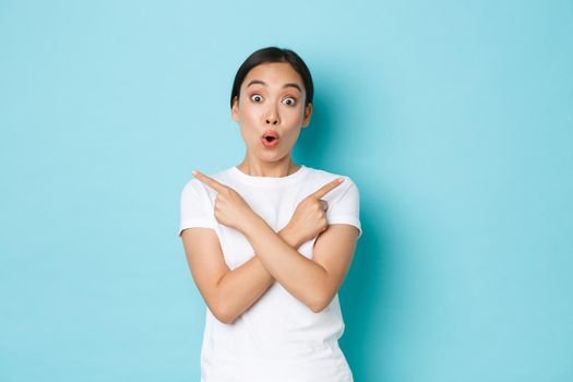 Amazed happy and surprised asian girl making announcement, woman pointing fingers sideways and empty spaces for your banner, looking astonished and cheerful, showing way, blue background.