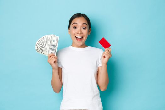 Shopping, money and finance concept. Amused asian girl in white casual t-shirt gasping, found out awesome prices, discount offers in shop, holding both credit card and cash, light-blue background.