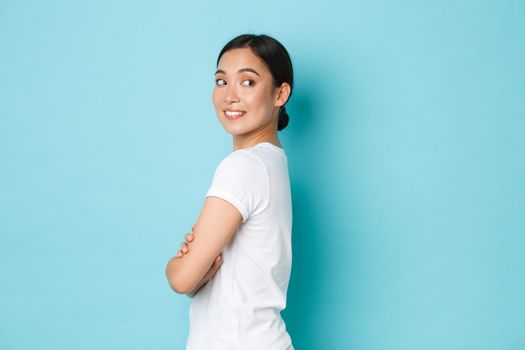 Coquettish pretty asian girl in white t-shirt standing in profile and turning right with curious smiling face, looking at something interesting, found good promo offer, standing blue background.