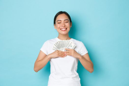 Shopping, money and finance concept. Lucky and pleased, happy asian woman hugging cash with eyes closed and broad satisfied smile, earn first salary, rejoicing over blue background.