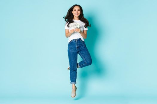 Fashion, beauty and lifestyle concept. Carefree beautiful asian girl looking upbeat, jumping dreamy and smiling while holding money, wasting cash over light blue background.