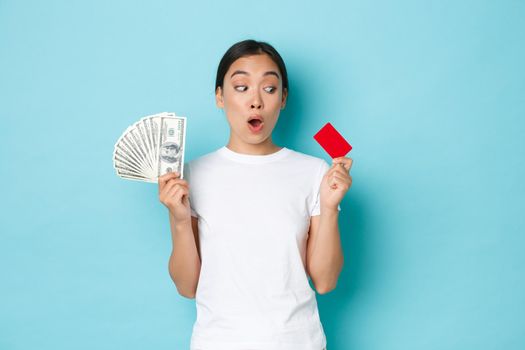 Shopping, money and finance concept. Amazed beautiful asian girl in white t-shirt gasping astonished and looking at credit card while holding cash in other hand, prefer contactless payment.