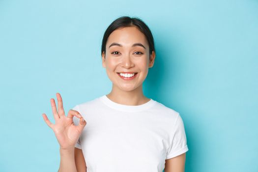 Close-up of pretty satisfied smiling asian girl in white t-shirt, showing recommendation gesture, okay sign and nod in approval, compliment choice, agree with you, standing light blue background.