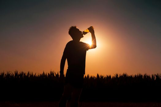 Young man drinks refreshing water while exercising and running. Silhouette of an athlete with a bottle of water on the background of the sun.