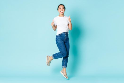 Fashion, beauty and lifestyle concept. Carefree smiling asian female looking joyful and upbeat, jumping over light blue background, showing thumbs-up in approval, recommend shop.