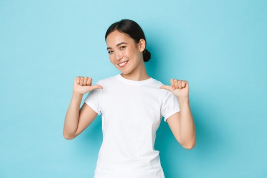Confident smiling asian woman in white t-shirt pointing at herself with proud, assertive expression, show-off, promote own abilities, searching for job, standing blue background.