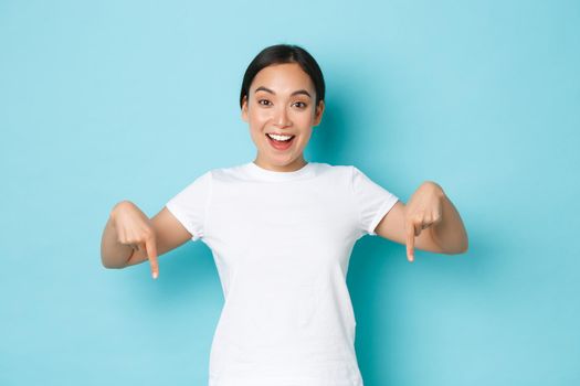 Cheerful young asian lady in white t-shirt pointing fingers down and smiling excited, looking upbeat while demontrating banner, offer special discount promo, standing blue background.